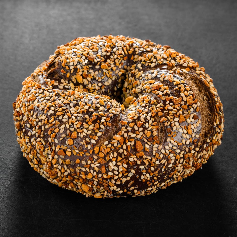 Dark bagel with a generous topping of sesame seeds and crispy garlic bits on a black surface.