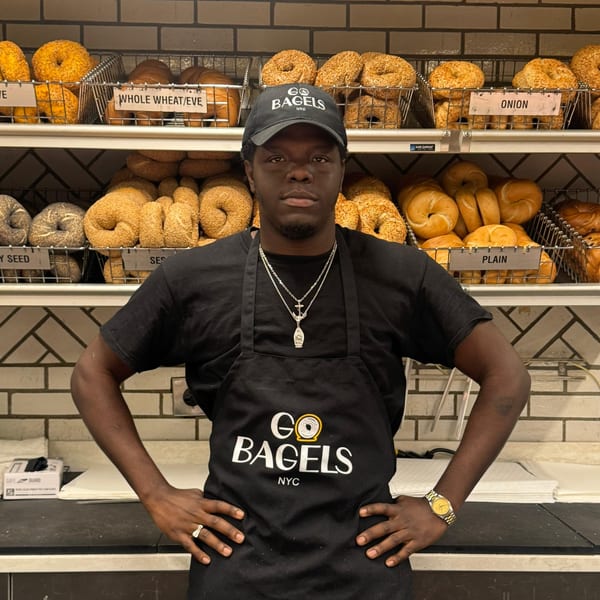 A Go Bagels NYC employee in a black apron and hat stands in front of shelves of bagels.