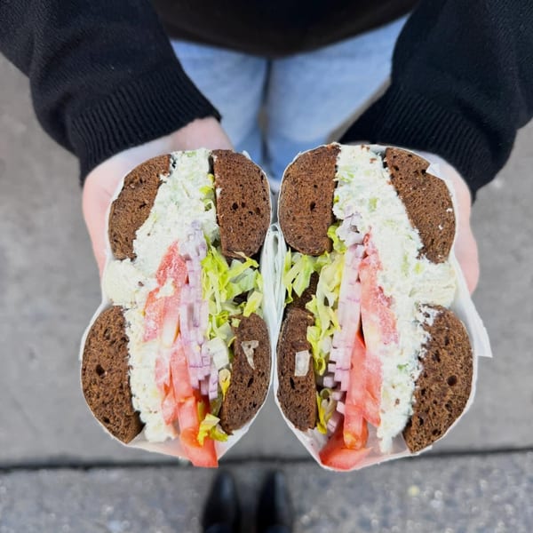 Hands holding a Go Bagels sandwich with dark bread, cream cheese, lettuce, tomatoes, and onions.
