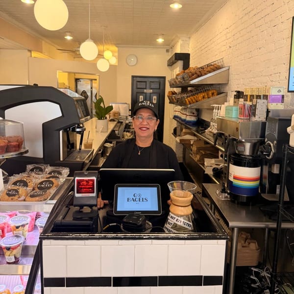A smiling woman in a "Go Bagels" hat stands behind a counter with a POS system and pastries.