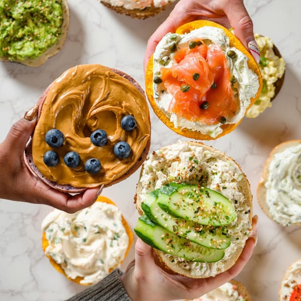 Hands holding Go Bagels with various toppings: peanut butter and blueberries, lox and capers, and cucumber.