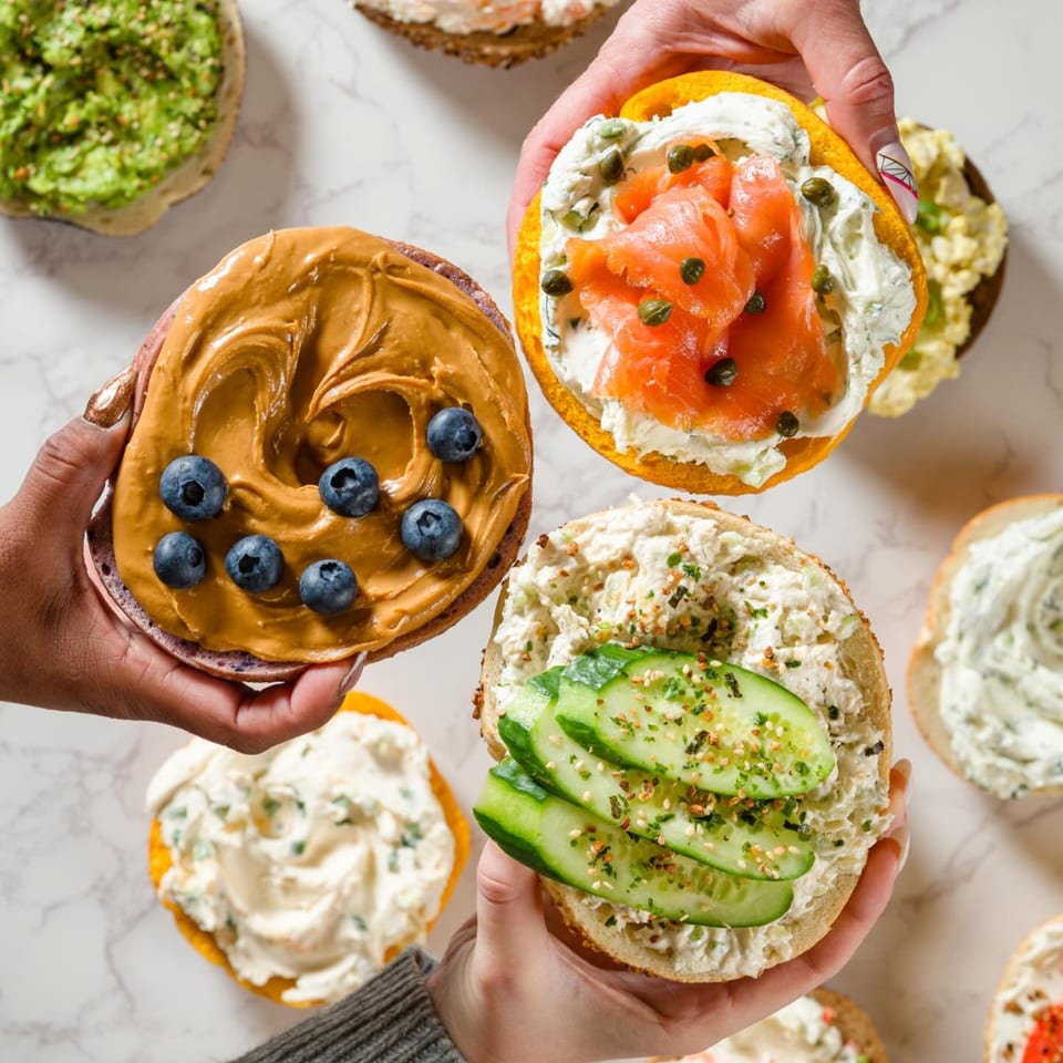 Hands holding Go Bagels with various toppings: peanut butter and blueberries, lox and capers, and cucumber.