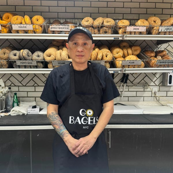 A man in a "Go Bagels NYC" apron stands in front of shelves filled with various bagels.