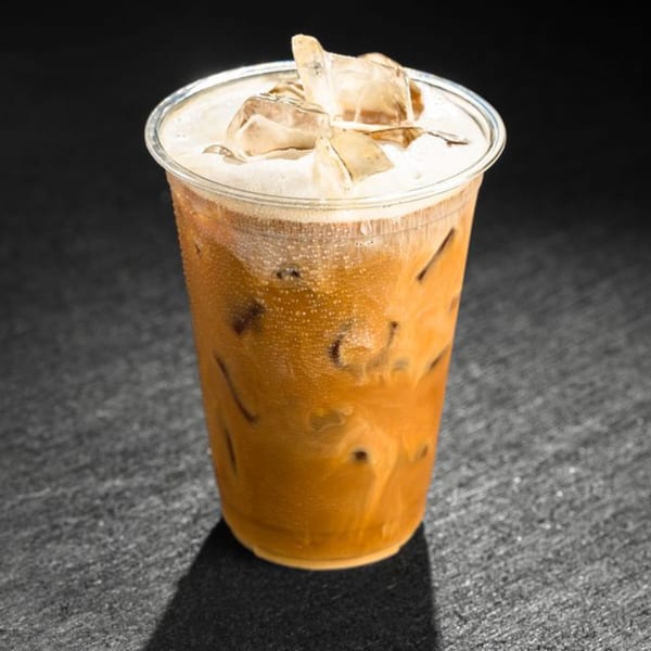 Iced coffee with foam and ice in a clear plastic cup on a dark, textured surface.