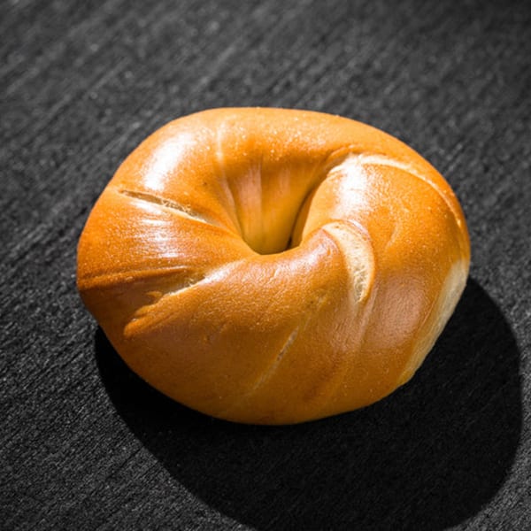 A single, golden-brown Go Bagel sits on a dark, textured surface, casting a shadow.