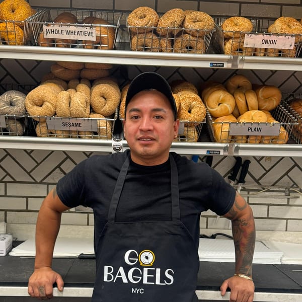 A man in a "Go Bagels NYC" apron stands in front of shelves filled with various bagels.