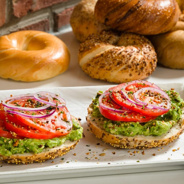 Two avocado and tomato bagels with red onion and seasoning, with plain bagels in the background.