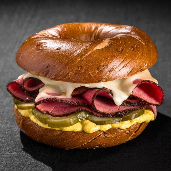Go Bagels pastrami bagel sandwich with melted cheese, pickles, and mustard on a dark surface.