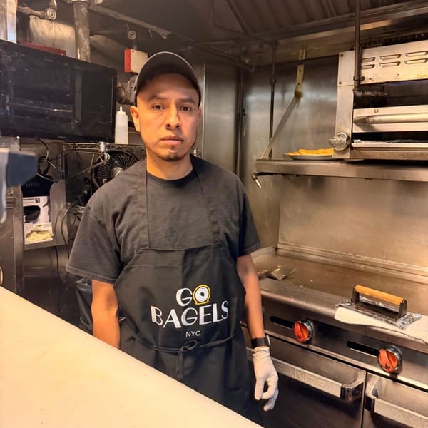 A Go Bagels NYC employee in a black apron and cap stands in a commercial kitchen.