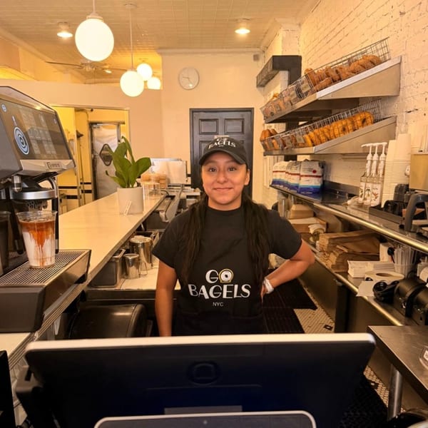 A Go Bagels NYC employee smiles behind the counter with bagels and coffee equipment.