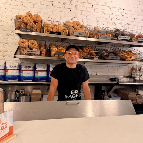 A smiling Go Bagels employee in orange glasses stands behind a counter with baskets of bagels.
