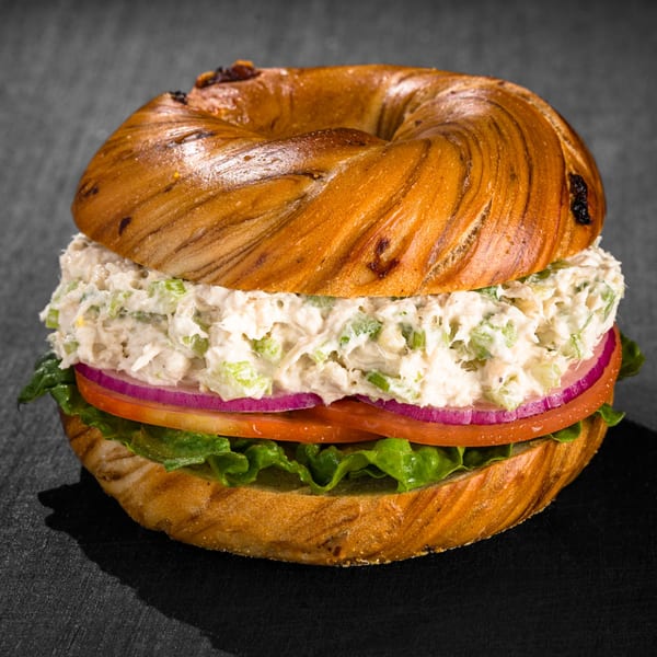 A Go Bagels chicken salad bagel sandwich with lettuce, tomato, and red onion on a dark surface.