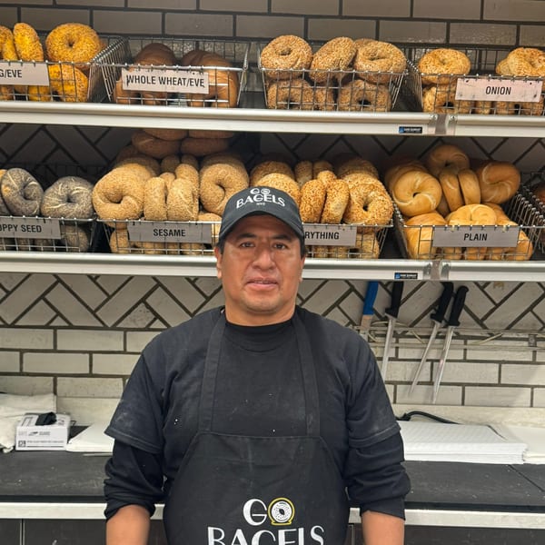 A man in a "Go Bagels" hat and apron stands in front of shelves of various bagels.