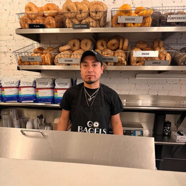 A man in a "Go Bagels" apron stands behind a counter with shelves of bagels and coffee bags.