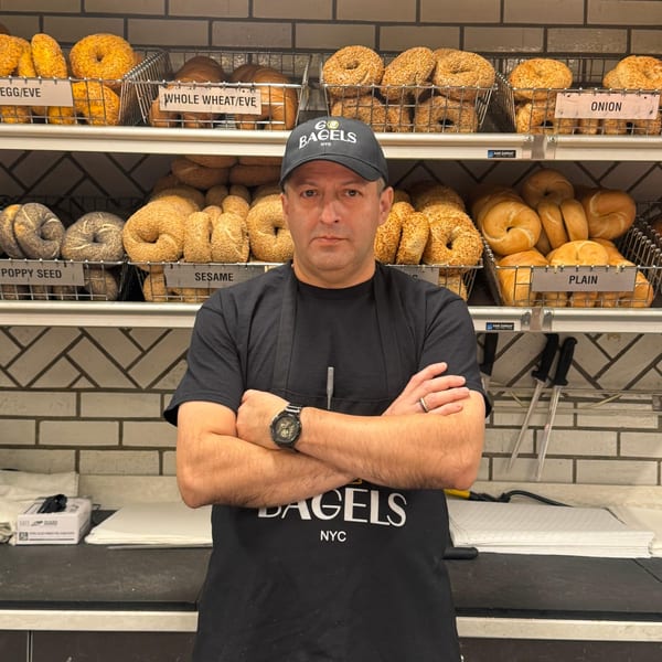 A man in a "Bagels NYC" hat and apron stands in front of shelves filled with bagels.