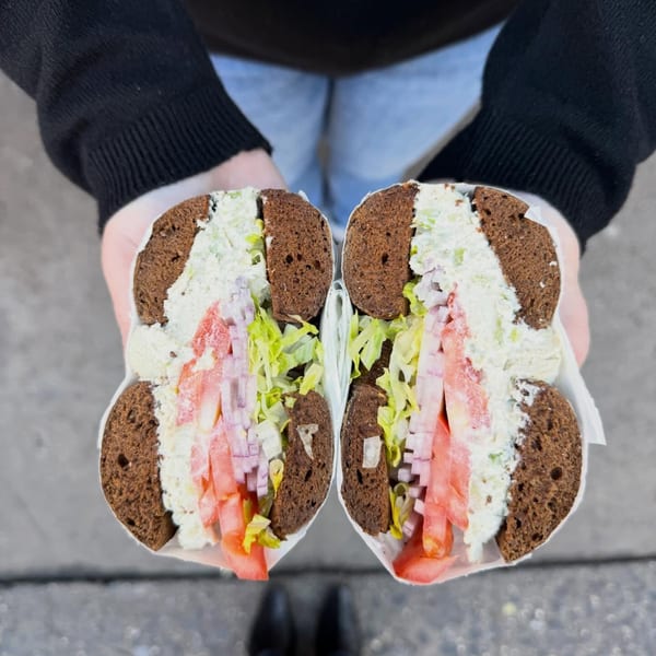 Hands holding a Go Bagels sandwich, sliced in half, revealing cream cheese, ham, lettuce, and tomatoes.
