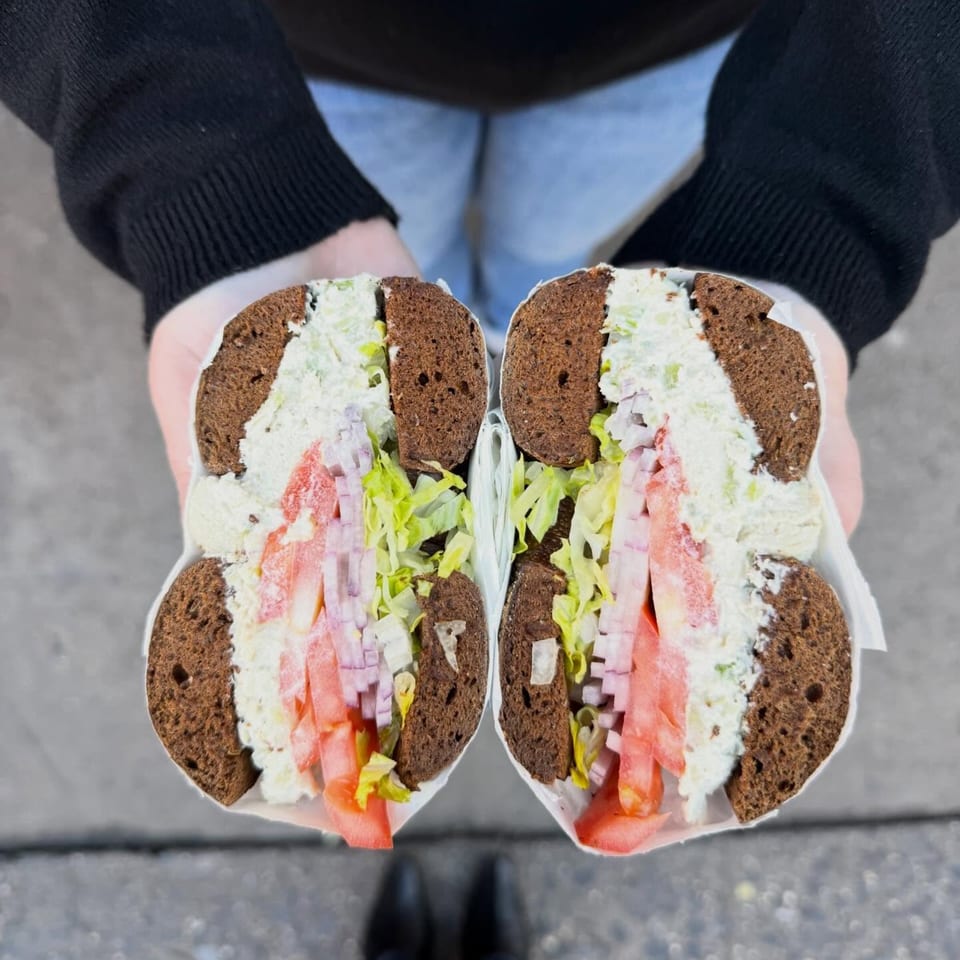 Hands holding a Go Bagels sandwich, sliced in half, revealing cream cheese, ham, lettuce, and tomatoes.