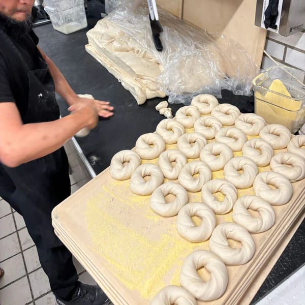 A baker shaping bagel dough by hand, with many raw bagels resting on a wooden board.