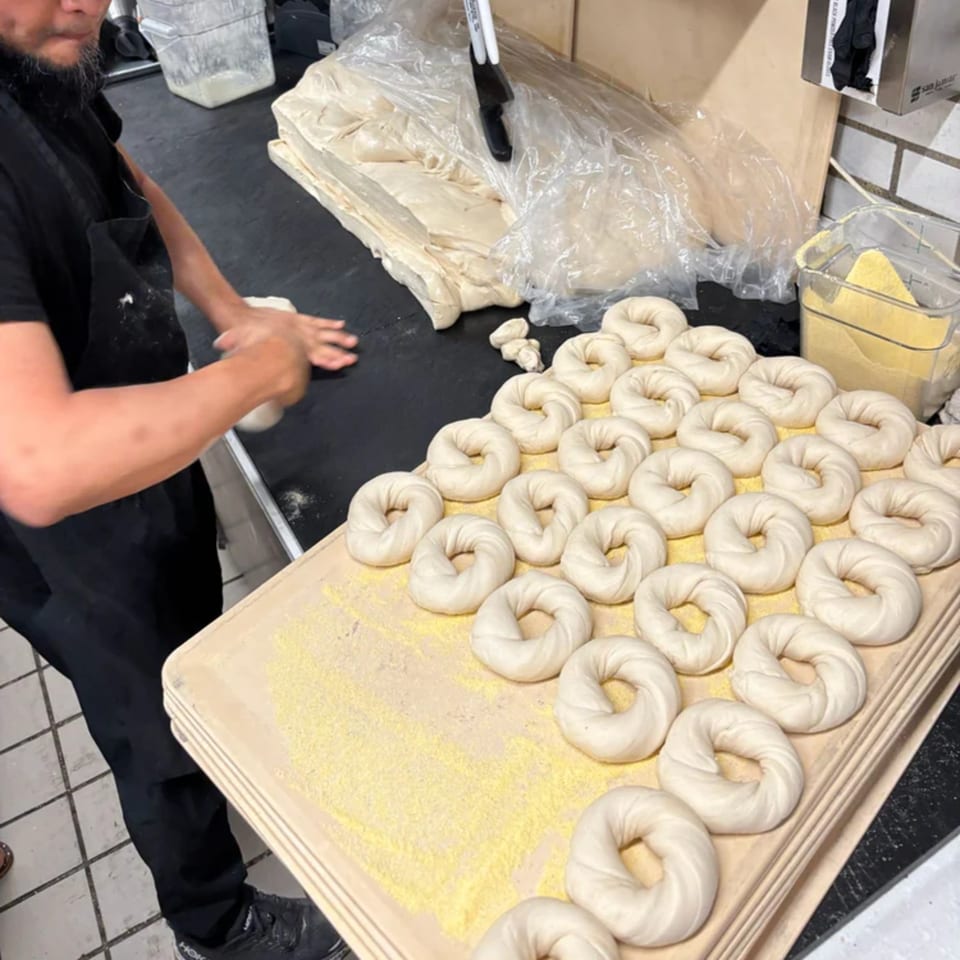 A baker shaping bagel dough by hand, with many raw bagels resting on a wooden board.