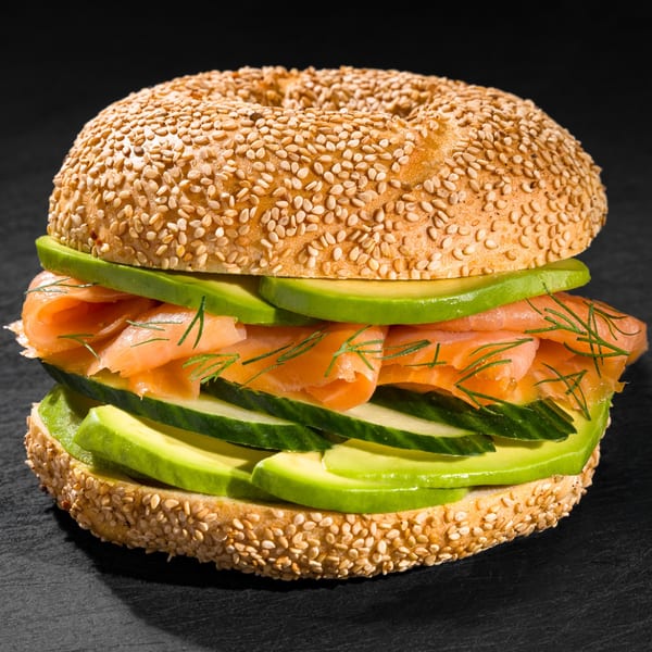 Sesame bagel with smoked salmon, avocado, cucumber, and dill on a dark surface.
