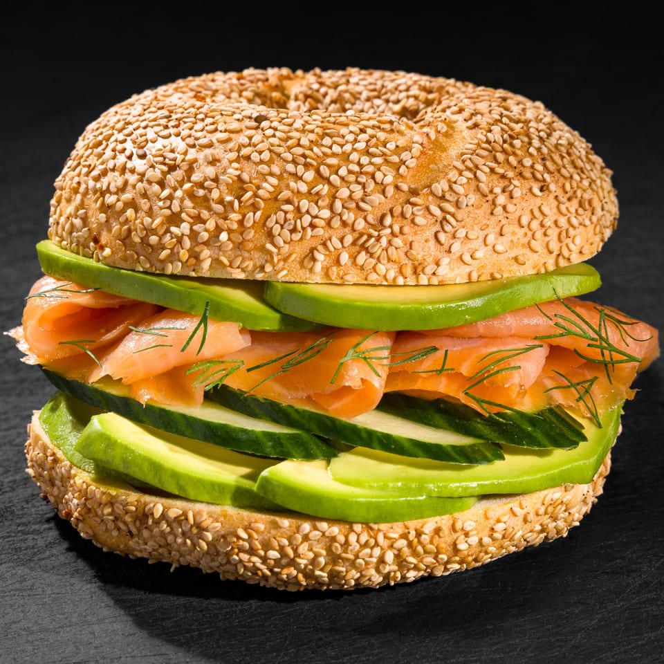 Sesame bagel with smoked salmon, avocado, cucumber, and dill on a dark surface.