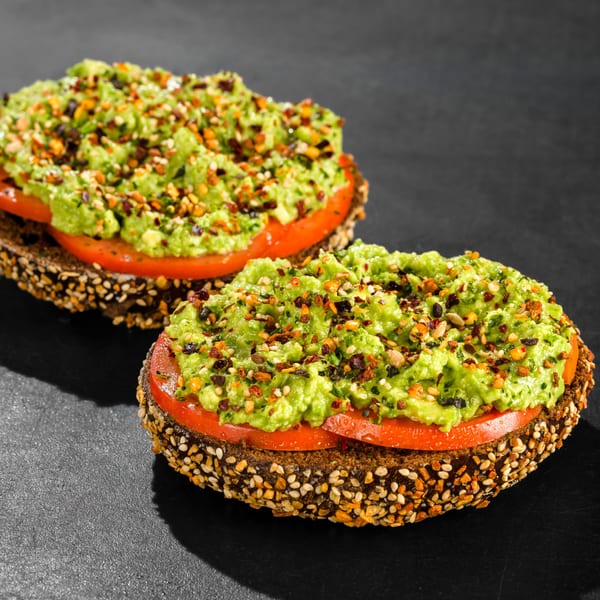 Two avocado toasts on seeded bagels with sliced tomatoes and everything seasoning on a dark surface.
